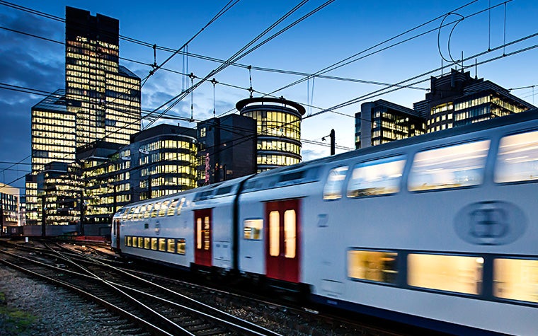 A white NMBS train traveling in the evening. Skyscrapers with lights on are in the background