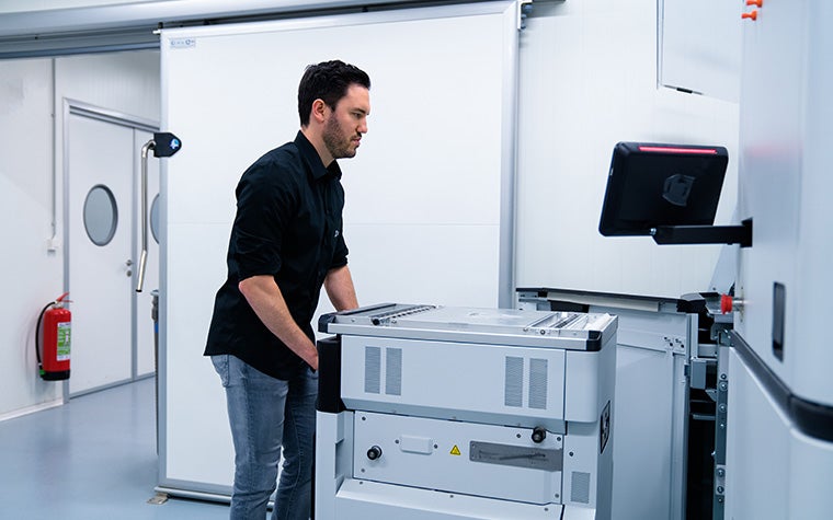 白い部屋に黒いTシャツと青いジーンズを着た男。部屋の中には3Dプリンターがあり、彼はプリンターのモニターを見ている。