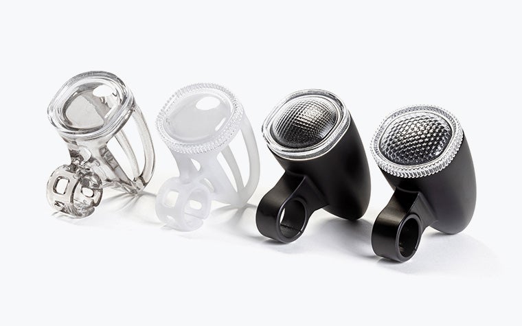 Four 3D-printed bicycle light housings made with VersaClear displayed in a row, showing different shades, from transparent and white prototypes to finished black units with textured reflective lenses.