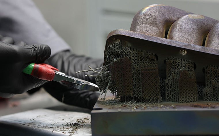 手袋をはめた人がペンチを持ち、3Dプリントされた部品から金属のサポート構造を取り除こうとしている。