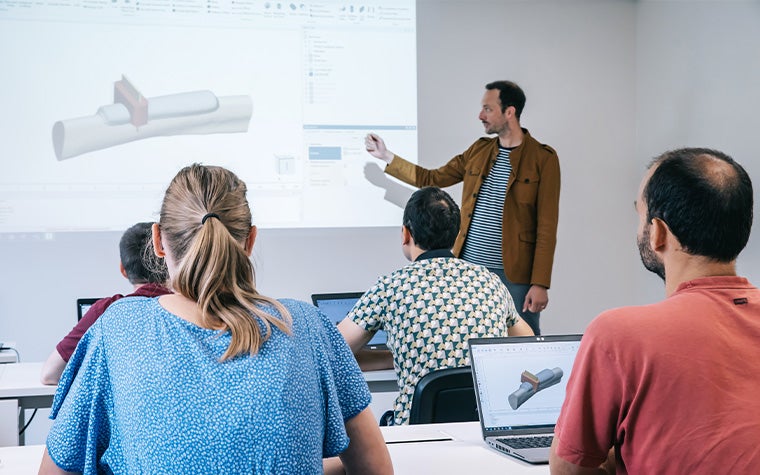 A man teaching a course to a room of people in front of a projection of a 3D rendered part.