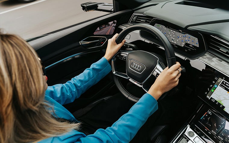 A woman driving an Audi car. An exterior/shoulder view of the car shows the dashboard, satnav, and the woman holding the steering wheel