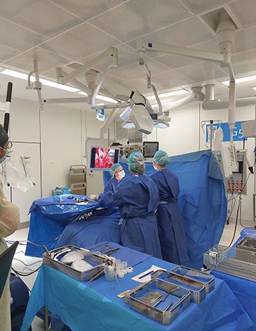 An operating room with a patient laying on the operating table and many surgical robotics hovering above them. Two surgeons stand on either side of the table. In the forefront, a desktop computer displays a visual of the patient's anatomy. In the background, a screen is hanging from the ceiling with the same visual. One of the surgeons is looking at this screen.