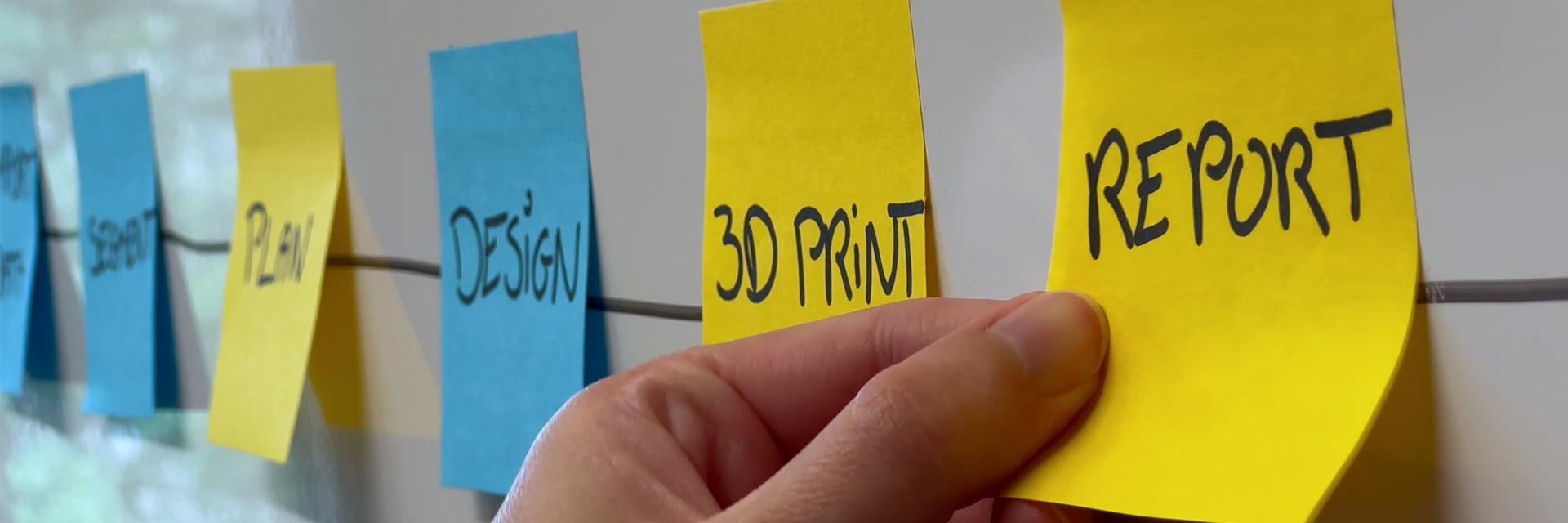 Hand placing a yellow sticky note labeled “REPORT” on a wall timeline alongside notes reading “PLAN,” “DESIGN,” and “3D PRINT.”