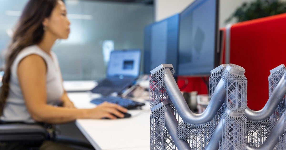 A complex, metal 3D-printed part with supports is in the foreground while a woman sitting at a desk looking as PC screen can be seen in the background.