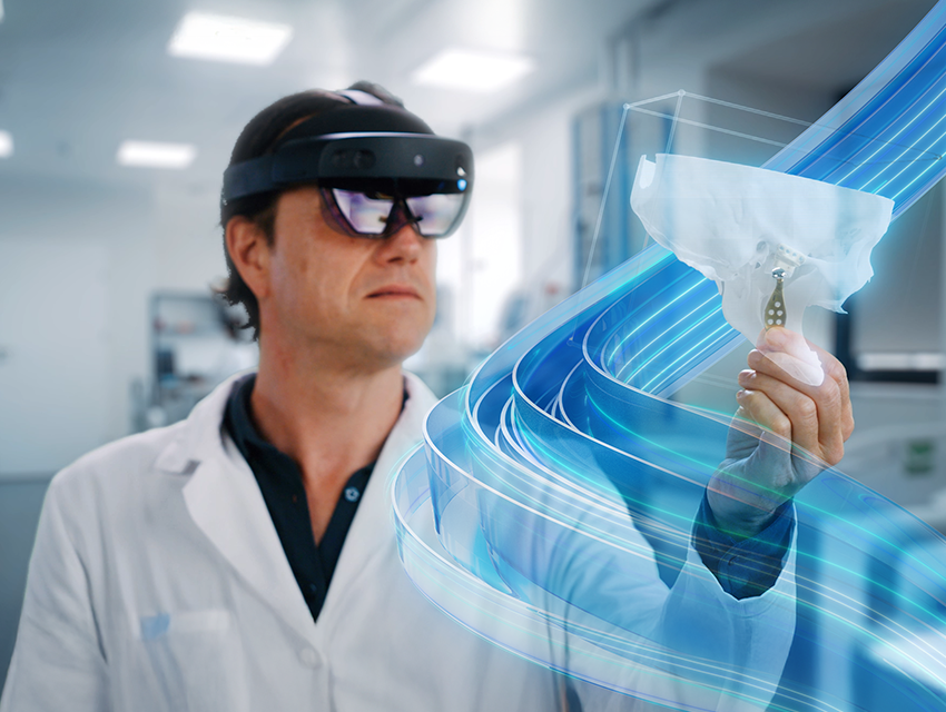 A surgeon wearing Microsoft Hololens augmented reality glasses and interacting with a virtual model of a patient's skull with a 3D-printed TMJ implant. The Mimics branded blue swirl is flowing around the model.