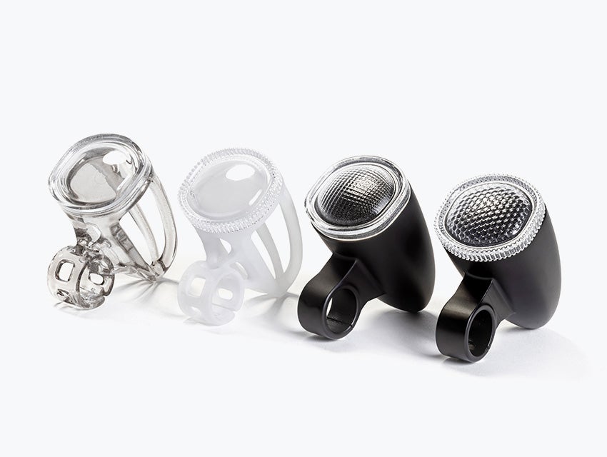 Four 3D-printed bicycle light housings made with VersaClear displayed in a row, showing different shades, from transparent and white prototypes to finished black units with textured reflective lenses.