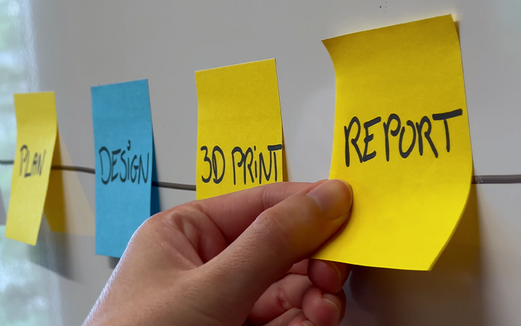 Hand placing a yellow sticky note labeled “REPORT” on a wall timeline alongside notes reading “PLAN,” “DESIGN,” and “3D PRINT.”