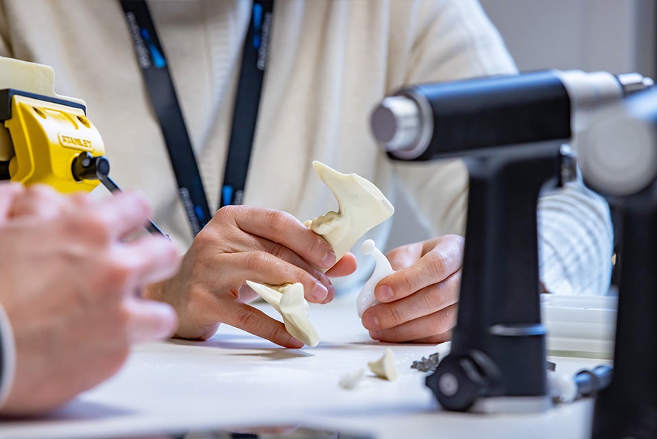 Participants handling anatomical bone models and surgical tools during a hands-on Materialise CMF Dry Bone training session, with powered surgical equipment on the table in the foreground.