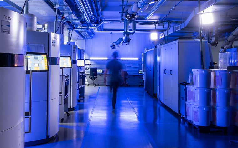 A person walking between rows of industrial selective laser sintering 3D printers in a machine park, dimly lit by a purple light.