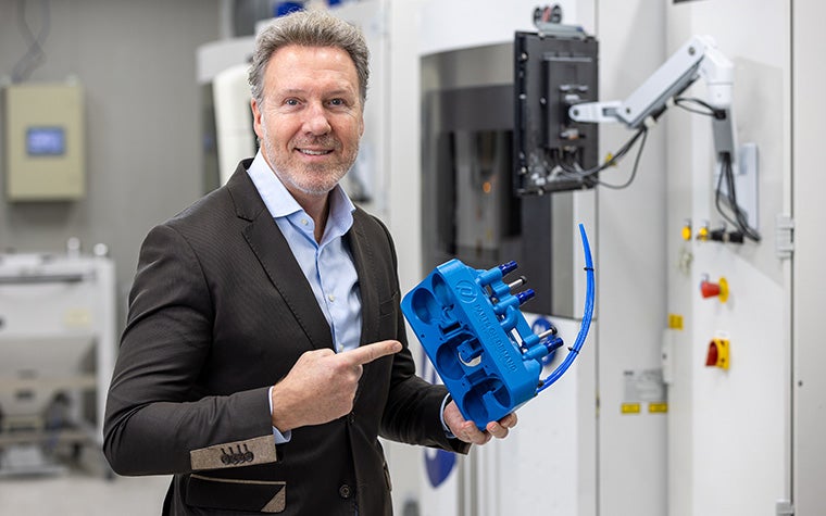 Frank Elbersen, Sales Engineer at Parts on Demand, pointing to a blue 3D-printed part in front of a 3D printer.
