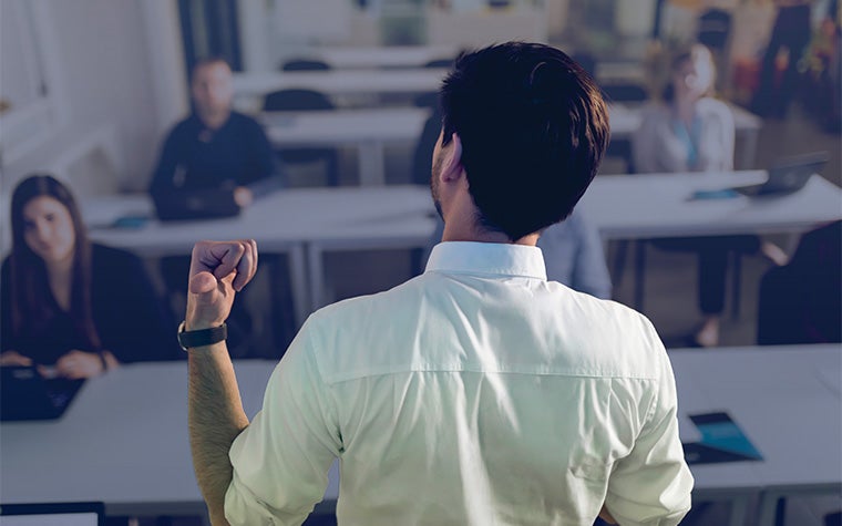 The back of a man raising his hand in front of a group of students looking at him.