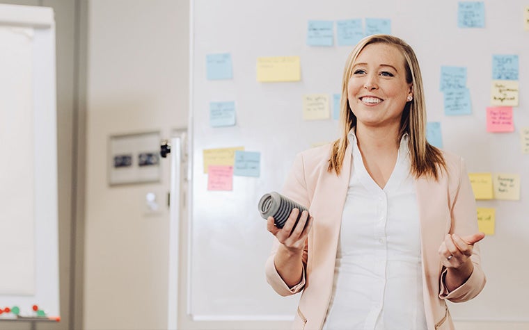 A blonde woman holding a 3D-printed object speaks in front of a whiteboard covered in post-it notes.