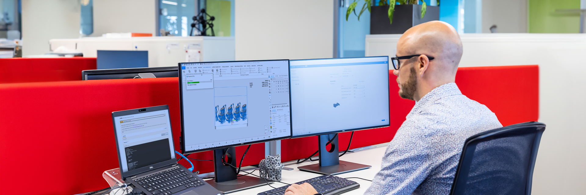 Man sitting at a desk with two computer screens and a laptop, working on a 3D printing build in Magics