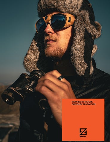 A man outdoors wearing a dark trapper hat and Zirkel's 3D-printed Peak Pioneer EXPLR Bison eyewear and holding binoculars.