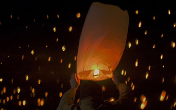 Person holding a lit paper lantern with many lanterns floating in the night sky in the background