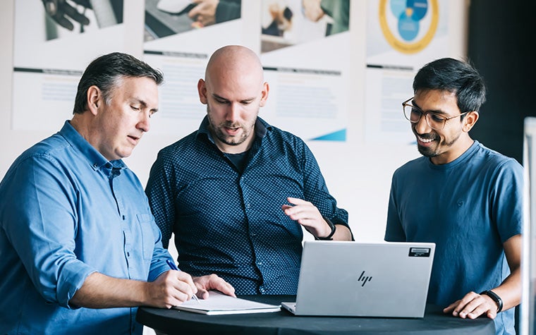 Two Materialise Academy trainees standing at a table with a laptop and speaking to the trainer. One of the men is taking notes