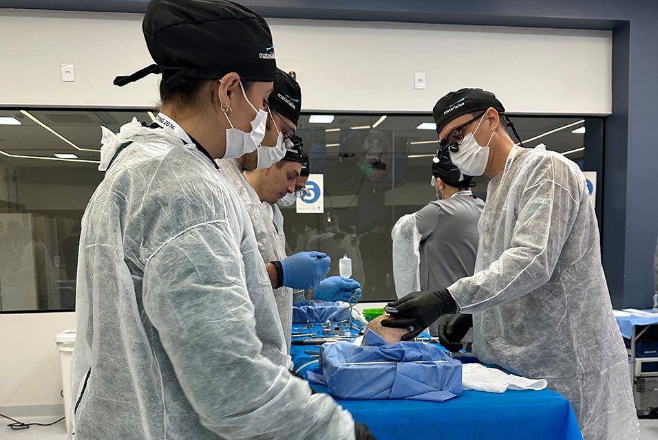 Medical professionals wearing protective gowns, masks, and surgical loupes while participating in a hands-on Materialise CMF Wet Lab training session around an operating table.