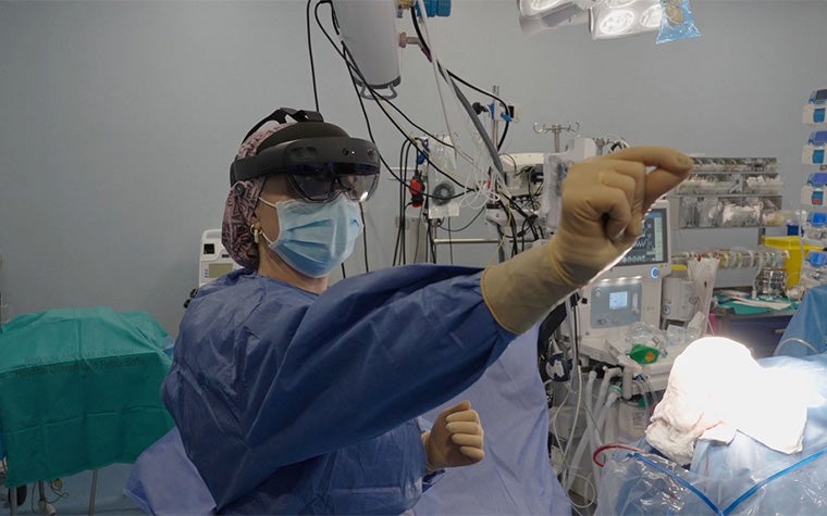 A surgeon wearing a head-mounted augmented reality Microsoft Hololens device gestures while working in an operating room with medical equipment in the background.