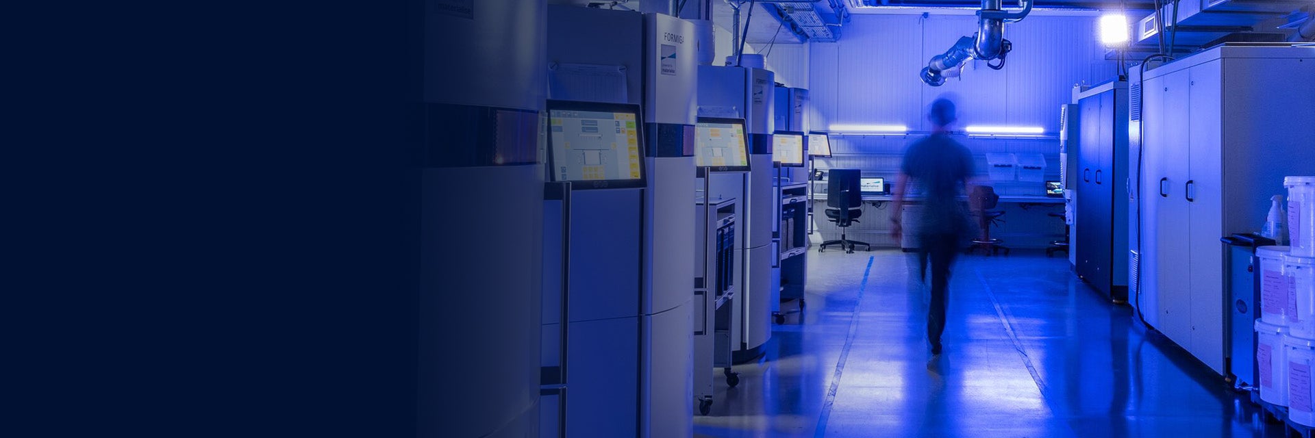 A person walking between rows of industrial selective laser sintering 3D printers in a machine park, dimly lit by a purple light.