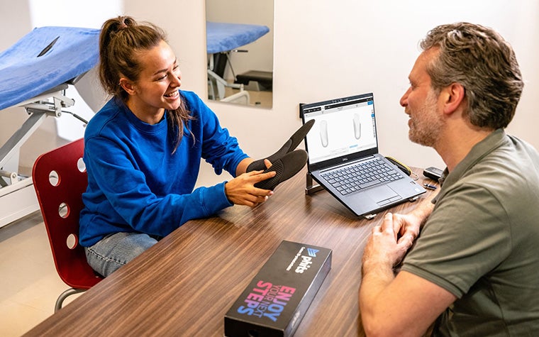 A woman holding a pair of custom-made phits orthotics sitting at a desk in a clinic opposite a male podiatrist.
