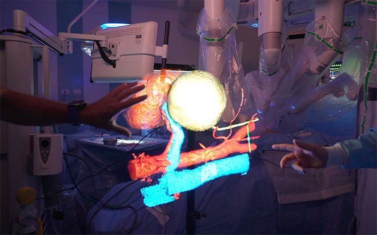 A holographic 3D medical visualization showing anatomical structures in red, blue, and yellow, with two people’s hands interacting with the floating image in a surgical setting.