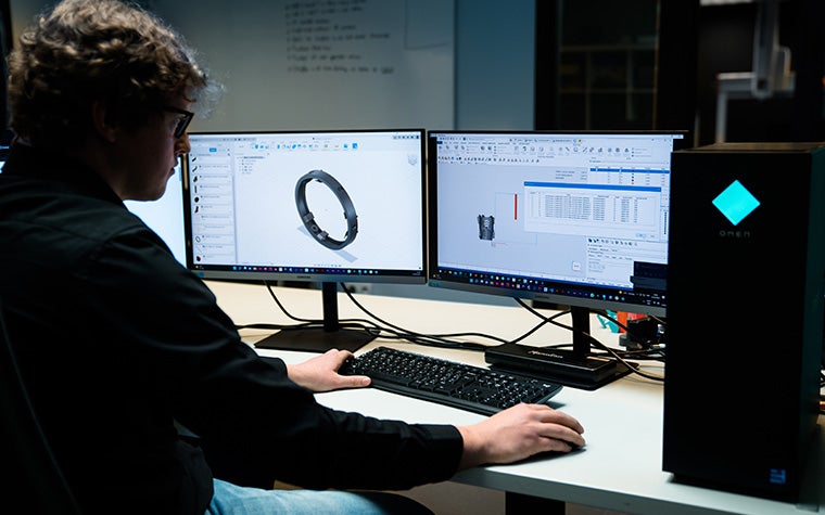 2つのスクリーンがあるデスクに座る男性。モニターには、Nesterモジュールにネストされた3Dモデルが表示されている。