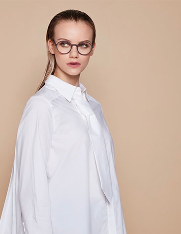 Weibliches Model in Weiß, das eine dunkle Brillenfassung von BAARS SELASI trägt und aus der Kamera schaut.