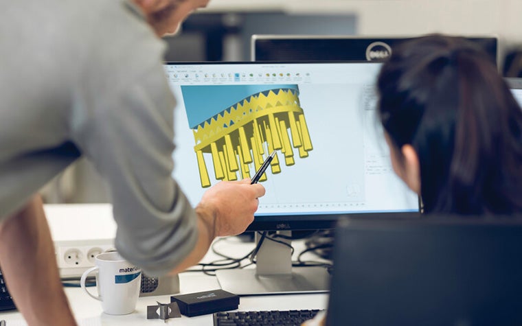 A man standing next to a woman sitting at a desk, using a pen to point to a 3D design in Magics software on a computer screen