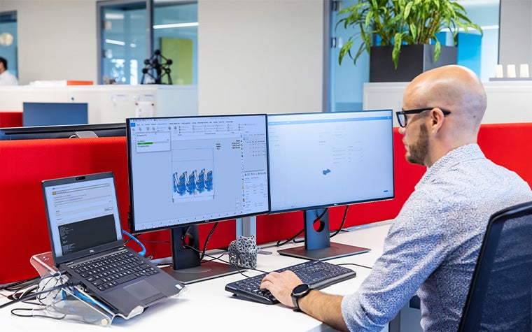 2つのコンピューター画面とノートパソコンが置かれた机に座り、Magicsで3Dプリントの製作に取り組む男性。