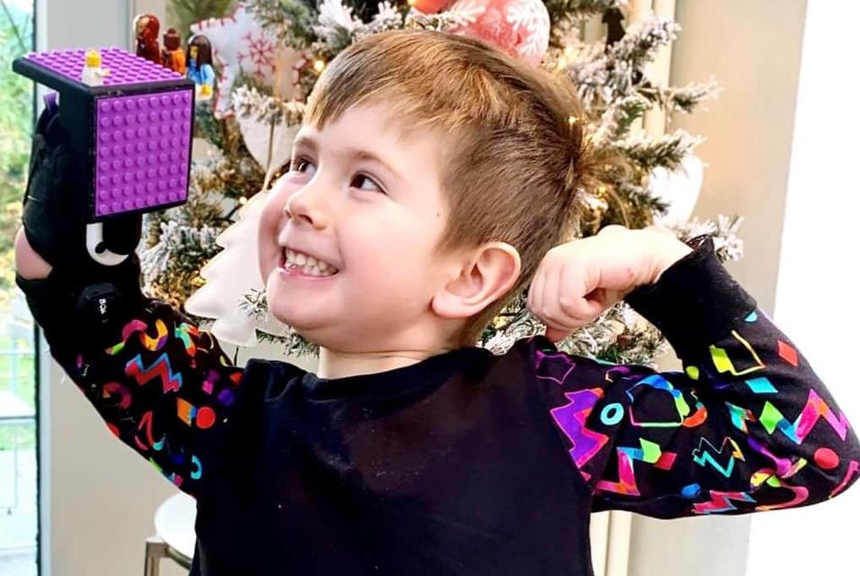 A smiling young boy wearing a Mitt prosthetic arm fitted with a LEGO attachment strikes a strongman pose