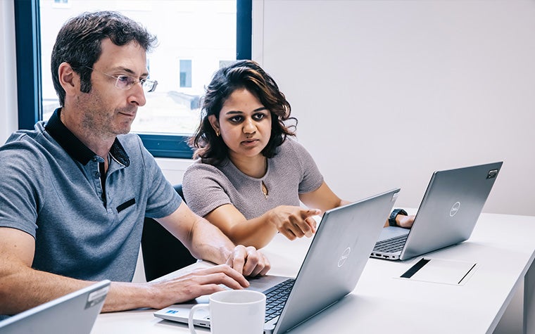 A man and a woman in a classroom sitting at a desk inspecting some information displayed on a laptop. The woman is pointing at the laptop 
