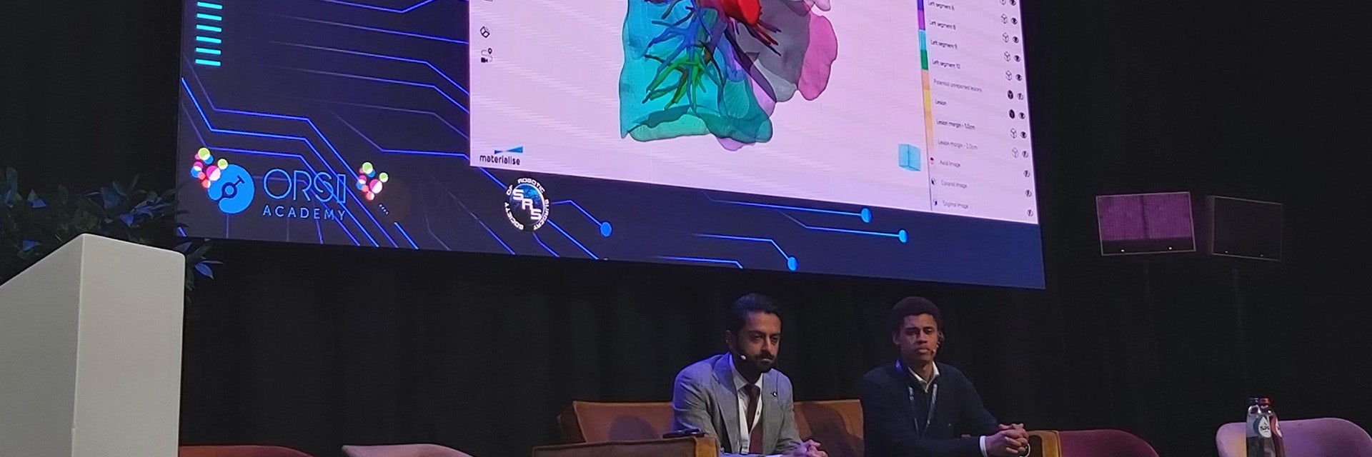 Two men sitting on a couch at Orsi Live, with a live view of a surgery and a surgical plan in Materialise Mimics Viewer displayed on a large screen behind them