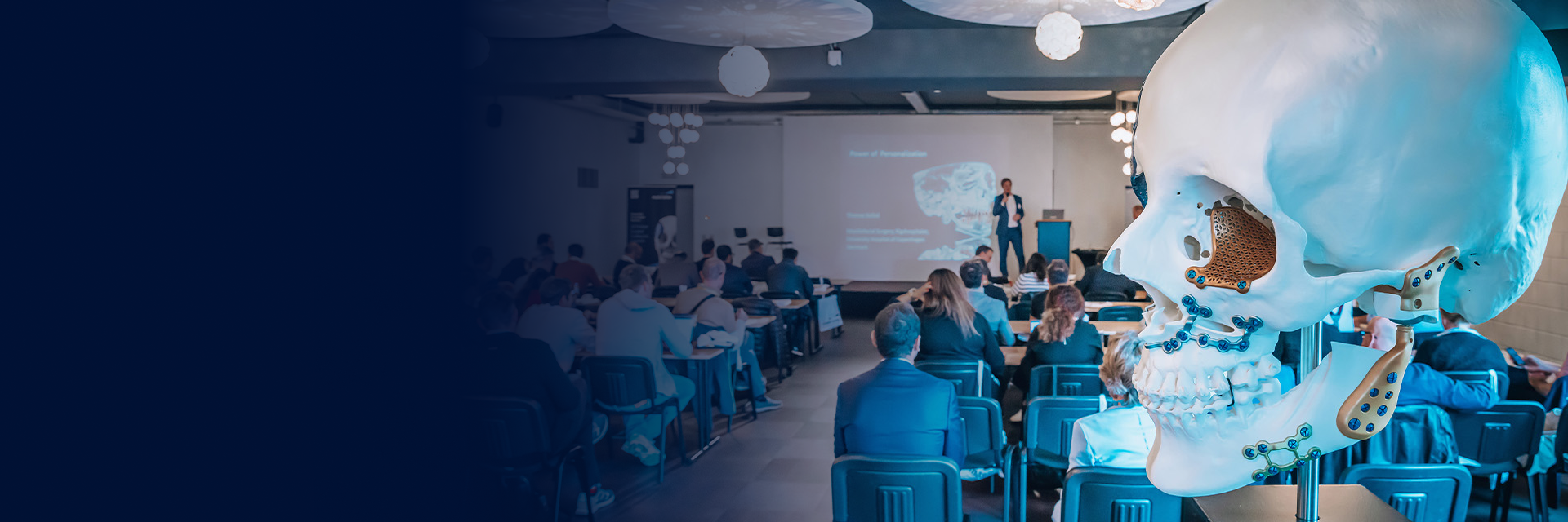 A large skull model with personalized implants attached in a meeting room with a presenter standing in front of a screen and event attendees sitting in seats at tables