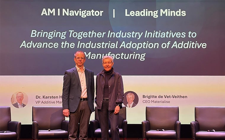 Siemens VP of Additive Manufacturing Dr. Karsten Heuser and Materialise CEO Brigitte De Vet standing on a stage in front of chairs, with a screen behind them reading "AM I Navigator | Leading Minds: Bringing Together Industry Initiatives to Advance the Industrial Adoption of Additive Manufacturing".