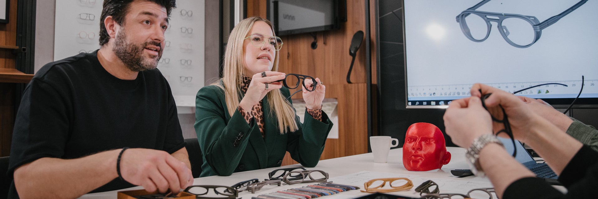 Persone sedute a un tavolo che parlano e osservano montature per occhiali stampate in 3D. Sullo sfondo si vede uno schermo con un progetto di montatura per occhiali