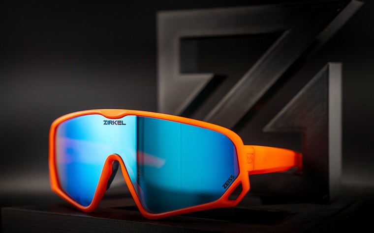 Zirkel's bright orange 3D-printed Teddy Grizz eyewear with a blue visor sitting against a dark grey and black background.