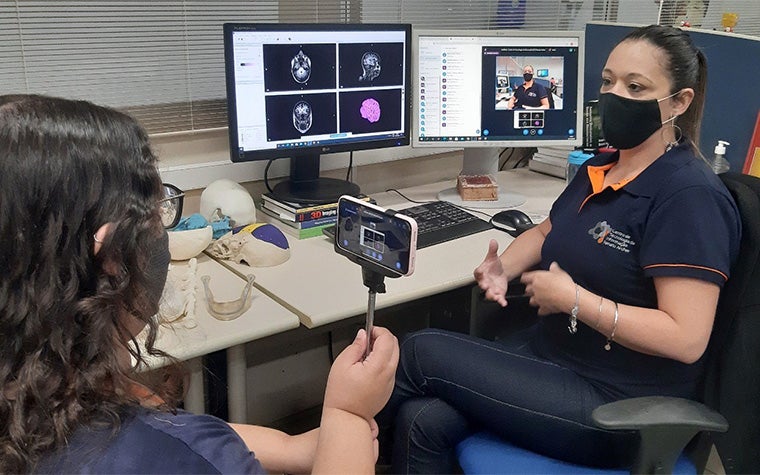 Woman recording Amanda Nunes with a mobile phone while she sits at a desk with anatomy software on open computer monitors