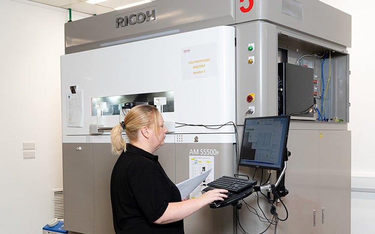 A Ricoh 3D production operator reviews a computer screen next to a 3D printer while holding a stack of papers