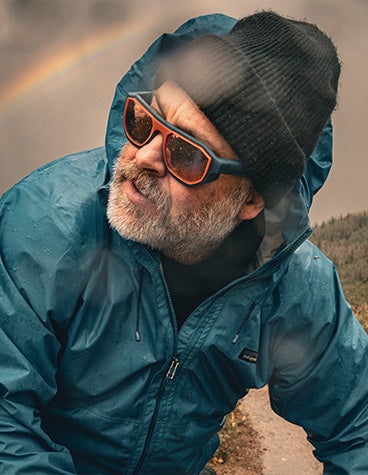 A man outdoors in rainy weather wearing a dark beanie, a blue windbreaker, and Zirkel's 3D-printed Sawtooth EXPLR x3 eyewear.