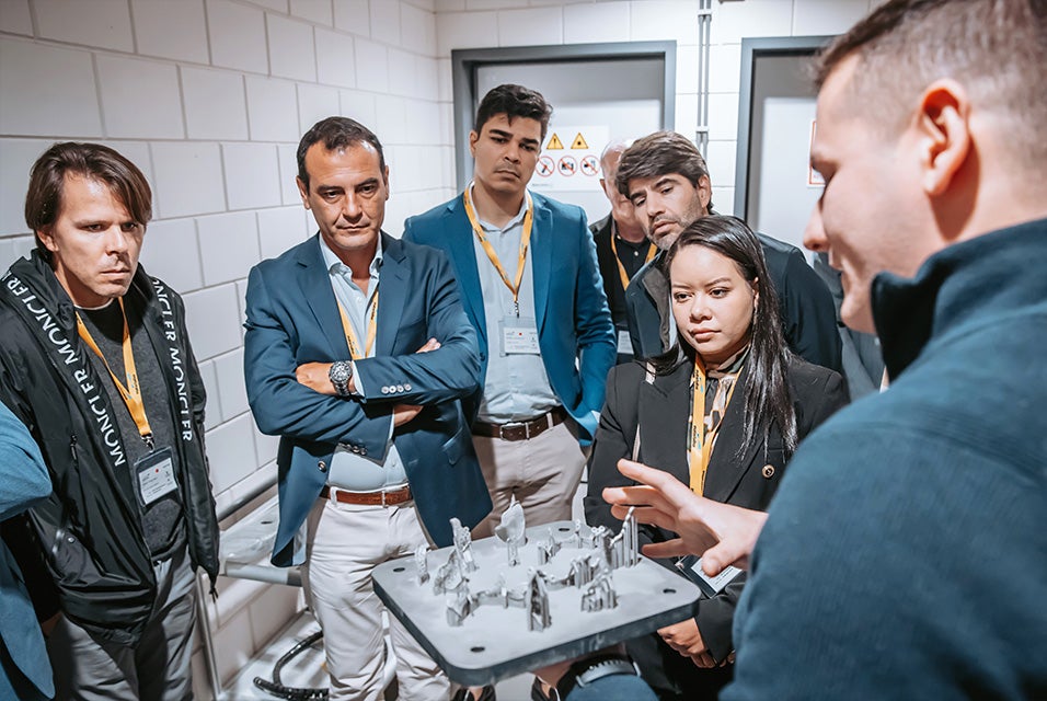 A group of Materialise CMF Innovation Summit attendees standing around a man holding a metal 3D printing build plate with various 3D-printed parts attached