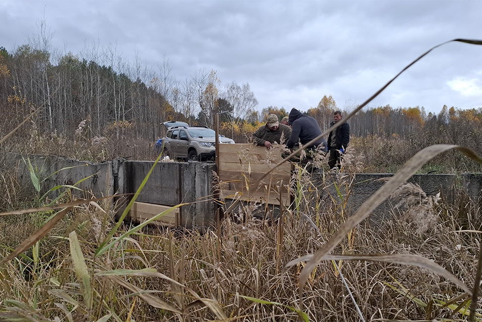 A group of men is putting up a wooden slider/sluice gate between a concrete wall in the Drevlyanskyi nature reserve. Dark brown foliage surrounds the gate and wall with a forest and a 4x4 vehicle in the background.