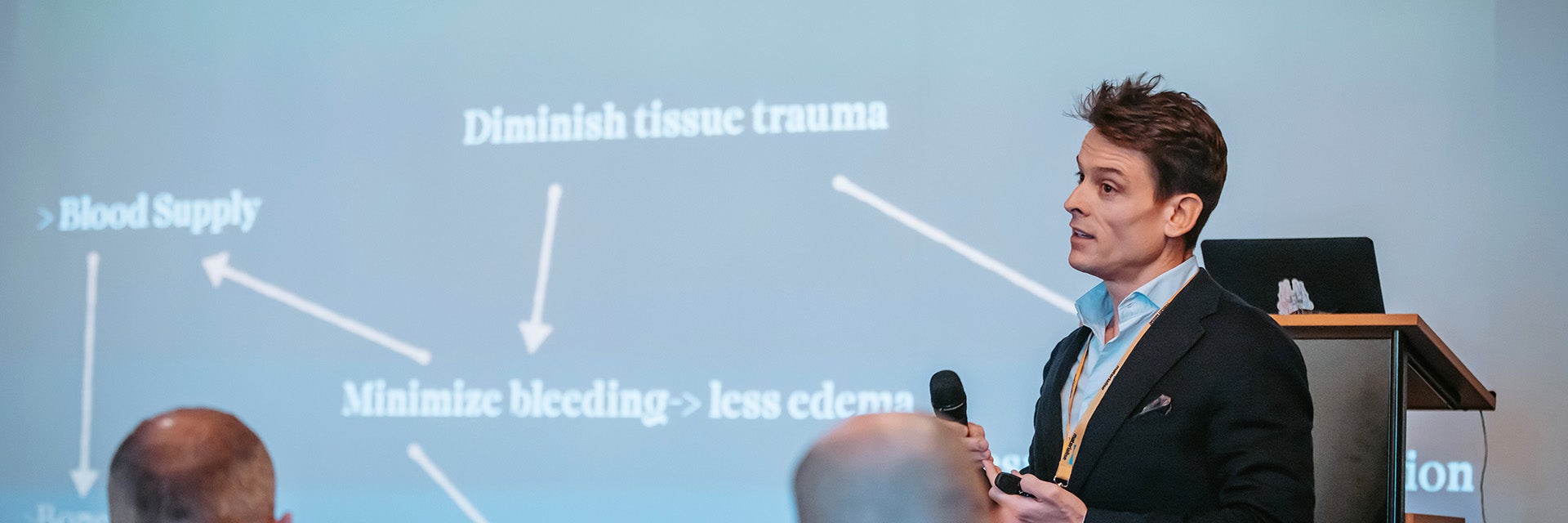 Dr. Javier Gutierrez speaking at the Materialise CMF Innovation Summit. The words "Diminish tissue trauma", "Blood supply", and "Minimize bleeding" show on the screen behind him, connected by arrows.