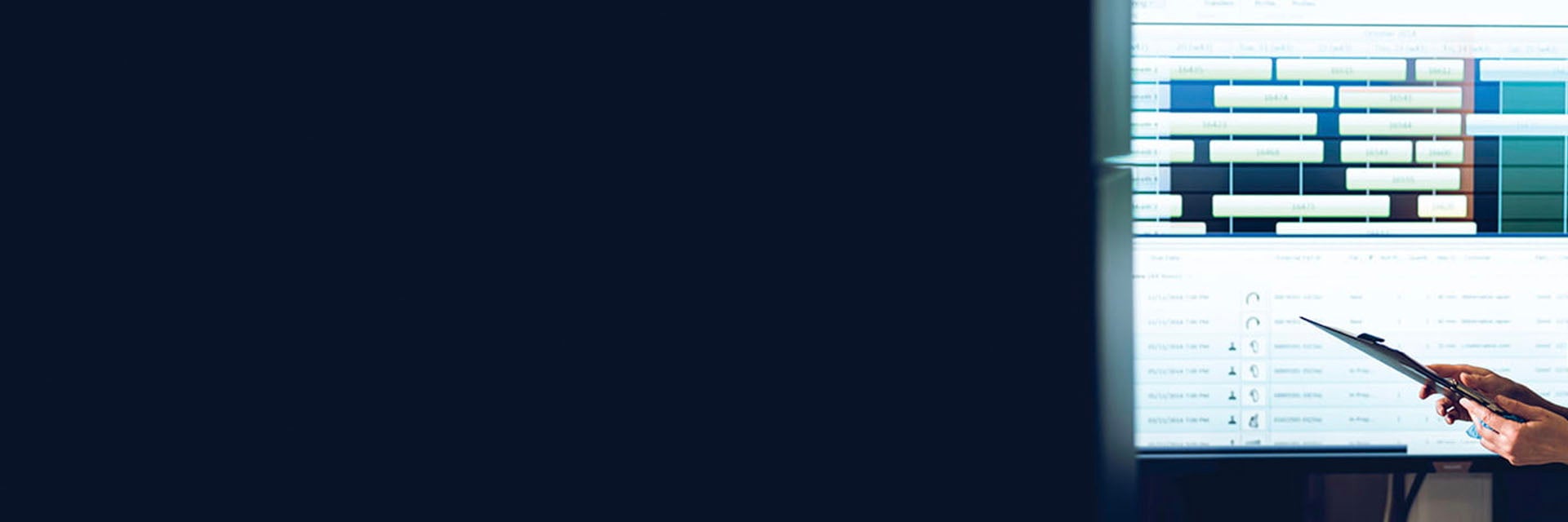 Person holding a clipboard, standing in front of a screen with production management software