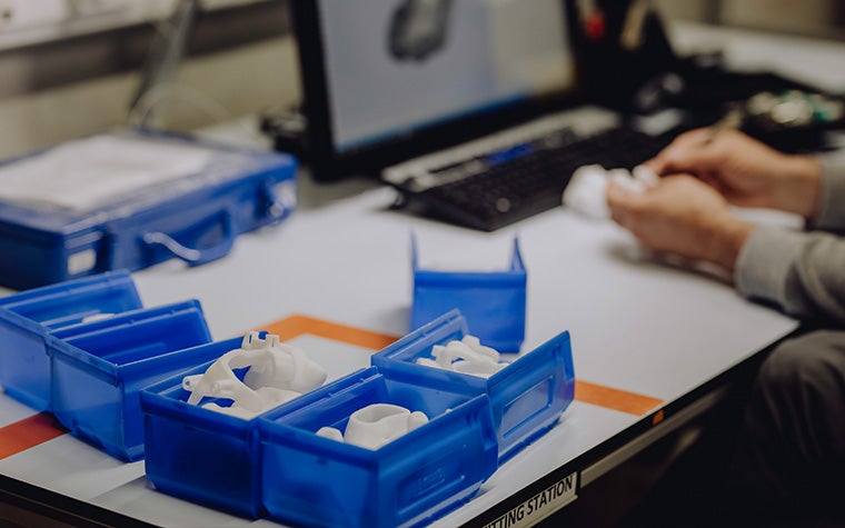 Seven blue containers with personalized medical 3D-printed implants and a man checking an implant in the background 