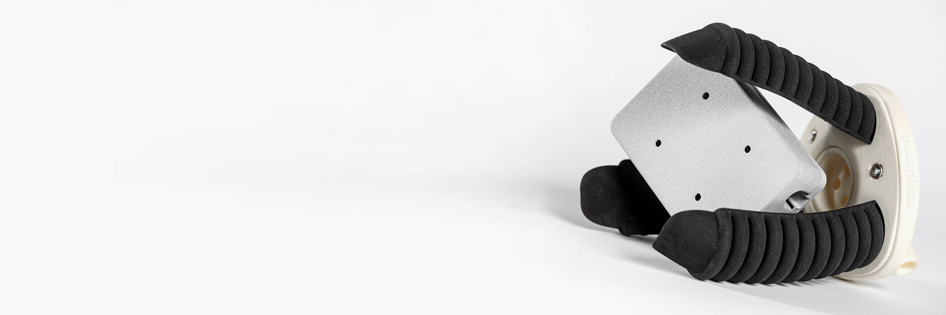 A gripper with a rigid, round base and three flexible pneumatic fingers, shaped like bellows. The gripper is shown lying on its side, and is not currently mounted on a robot. A small box-like part is shown in the gripper fingers as an example of common pick-and-place assembly line tasks.