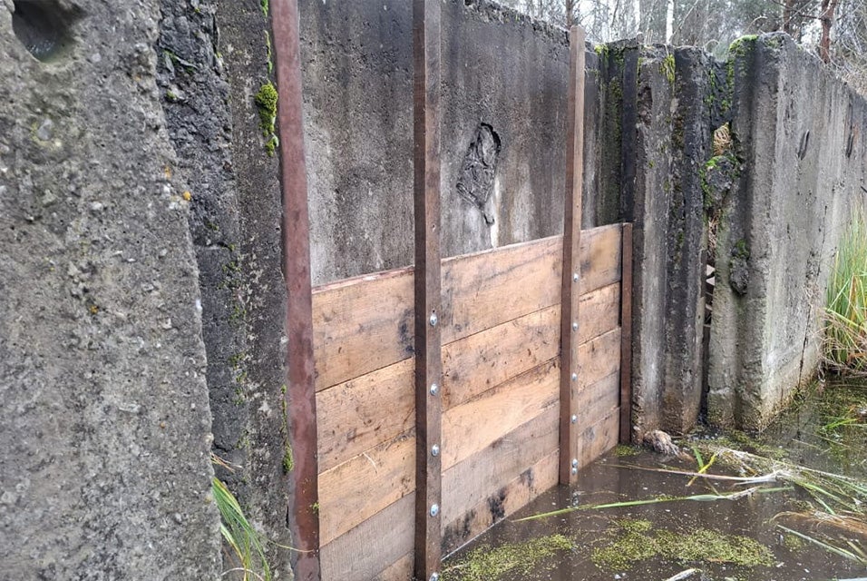 A wooden slider/sluice gate in between a concrete fence in the Drevlyanskyi nature reserve. The gate has four panels and is submerged in water.