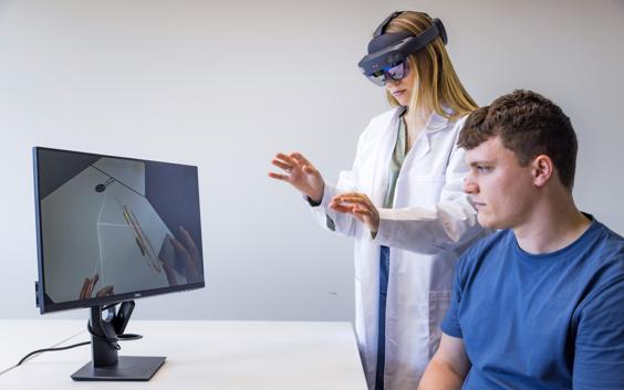 Ein Arzt trägt eine AR-Brille, während er virtuell Messungen vornimmt, während ein Mann den Vorgang auf einem Monitor beobachtet