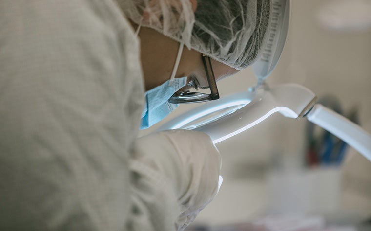 A medical professional/lab technican in white overalls inspecting a 3D-printed part under a magnifying glass