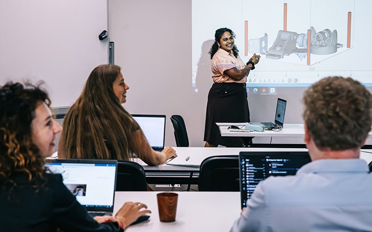 A Materialise trainer standing in front of a projector showing a 3D printing build in Materialise Magics. A group of student sit at tables in the foreground